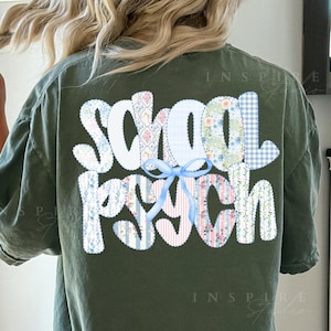 May include: Moss green t-shirt with the words "School Psych" in a patchwork design on the back. The patchwork includes floral, gingham, and striped patterns, with a light blue bow accent. The shirt is a comfortable, casual style.