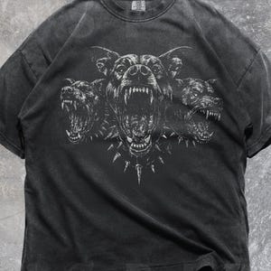 May include: Dark gray t-shirt featuring a three-headed dog graphic with open mouths and sharp teeth. The design includes a spiked collar. The shirt has a vintage, worn look. The word "Pepper" is at the bottom left, and "INSPIRE Studio" is at the bottom right.