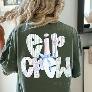 May include: Moss green t-shirt with the text "Eip Crew" in a floral design with a light blue bow. The shirt is a comfortable, casual style.
