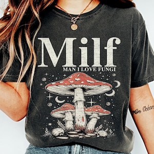 May include: A dark gray t-shirt with the text "Milf Man I Love Fungi" and a graphic of three mushrooms with red caps and white stems. The mushrooms are surrounded by stars and moons.