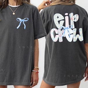 May include: Dark gray t-shirt with a light blue bow on the front and the words "Eir Crew" on the back. The text on the back is in a floral pattern with a light blue bow. The shirt is a comfortable, oversized fit.