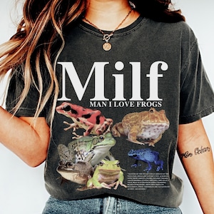 May include: A dark gray t-shirt with the text "Milf Man I Love Frogs" printed in white. The shirt features a colorful graphic of frogs.