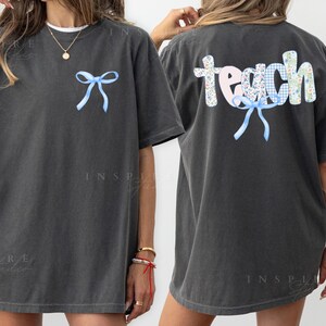 May include: Dark gray t-shirt with a light blue bow on the front and the word "teach" on the back. The word "teach" is made up of floral and gingham patterns. The shirt is a casual, oversized style.