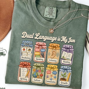 May include: A moss green crew neck t-shirt featuring the text "Dual Language is My Jam" above eight illustrated jars. Each jar contains icons representing vocabulary, speaking, listening, reading, writing, culture, communication, and growth.