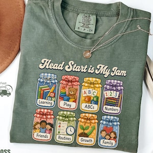 May include: A moss green crewneck t-shirt featuring the text Head Start is My Jam above eight illustrated jars. Each jar contains a different theme: Learning, Play, ABCs, Numbers, Friends, Routines, Growth, and Family, with colorful graphics inside.