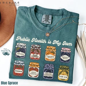 K&ouml;nnte beinhalten: Blaues Fichten-T-Shirt mit dem Aufdruck "Public Health is My Jam" und Illustrationen von Marmeladengl&auml;sern. Jedes Glas hat ein anderes Etikett, darunter "Advocacy" und "Health Promotion". Eine goldene Halskette liegt &uuml;ber dem Shirt.