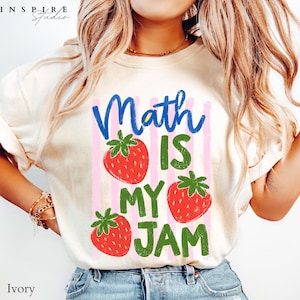 May include: An ivory short-sleeve t-shirt featuring a graphic print with the blue text "Math" and green text "IS MY JAM". The design includes three red strawberries with green leaves and vertical light pink stripes in the background.
