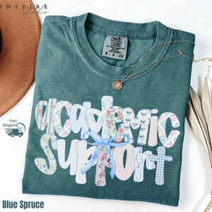 May include: A blue spruce Comfort Colors t-shirt with the words "Academic Support" in a patchwork design. The patchwork includes floral, bow, and striped patterns. A gold necklace with a pendant is on the shirt.