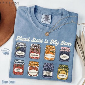May include: Blue jean colored t-shirt with the phrase "Head Start is My Jam" and illustrations of jam jars. Each jar has a different fruit and label, including "Cognitive" and "Resilience". A gold necklace is draped across the shirt.