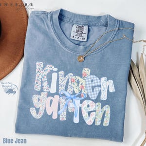 May include: A blue jean Comfort Colors t-shirt with the word "Kindergarten" in a patchwork design. The letters are made of floral, gingham, and striped fabrics. A light blue bow is under the word "garten". A gold necklace is draped across the shirt.