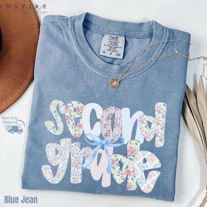 May include: A blue jean colored t-shirt with the words "Second Grade" in a floral and striped design. The shirt has a small blue bow and a gold necklace. The shirt is a size small.