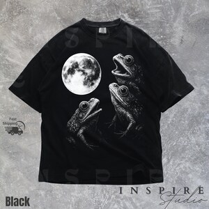K&ouml;nnte beinhalten: Schwarzes T-Shirt mit einer detaillierten Illustration von drei Fr&ouml;schen und einem Vollmond. Die Fr&ouml;sche sind in einer Gruppe, mit weit ge&ouml;ffneten Augen. Der Text "INSPIRE Studio" steht unten.