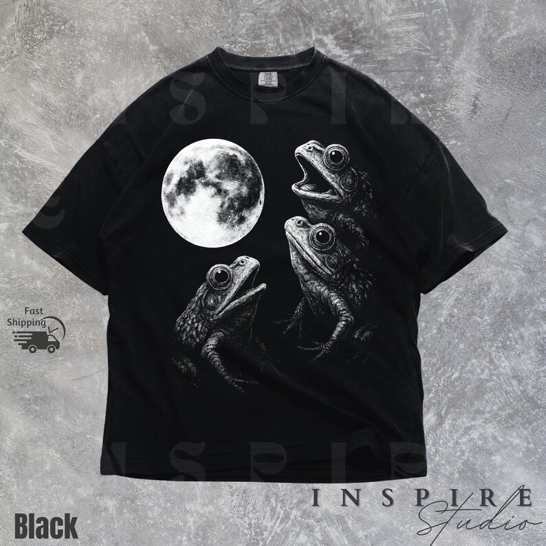 K&ouml;nnte beinhalten: Schwarzes T-Shirt mit einer detaillierten Illustration von drei Fr&ouml;schen und einem Vollmond. Die Fr&ouml;sche sind in einer Gruppe, mit weit ge&ouml;ffneten Augen. Der Text "INSPIRE Studio" steht unten.