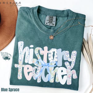 May include: A blue spruce Comfort Colors t-shirt with the words "history teacher" in white, floral, and gingham patterns. A light blue ribbon is tied below the word "teacher". A gold necklace with a pendant is on the shirt.