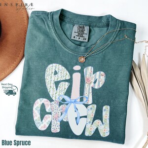 May include: A blue spruce Comfort Colors t-shirt with the text "Eip Crew" in a floral and striped design. A blue ribbon is tied at the bottom of the text. A gold necklace with a pendant is draped on the shirt.