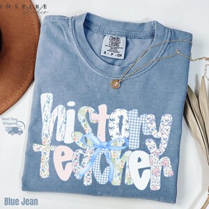 May include: A blue jean Comfort Colors t-shirt with the words "history teacher" in a patchwork design. The patchwork includes floral, gingham, and striped patterns. A gold necklace with a pendant is draped on the shirt.