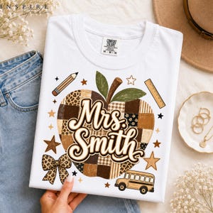 May include: A white t-shirt featuring a large apple graphic made of brown and tan patchwork patterns. The text "Mrs. Smith" is written in a brown script font across the apple. Surrounding the apple are a pencil, ruler, school bus, and bow.