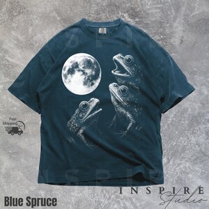 K&ouml;nnte beinhalten: Blau-Fichte-T-Shirt mit einer wei&szlig;en Illustration von drei Fr&ouml;schen und einem Vollmond. Die Fr&ouml;sche sind in einem detaillierten, skizzenartigen Stil gezeichnet. Der Text "Blue Spruce" befindet sich am unteren Rand des T-Shirts.