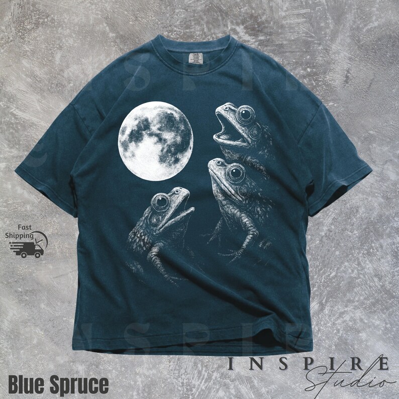 K&ouml;nnte beinhalten: Blau-Fichte-T-Shirt mit einer wei&szlig;en Illustration von drei Fr&ouml;schen und einem Vollmond. Die Fr&ouml;sche sind in einem detaillierten, skizzenartigen Stil gezeichnet. Der Text "Blue Spruce" befindet sich am unteren Rand des T-Shirts.