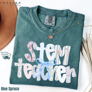 May include: Blue spruce t-shirt with the words "STEM Teacher" in a patchwork design with floral and striped patterns. A light blue bow accents the design. A gold necklace with a pendant rests on the shirt.
