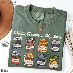 K&ouml;nnte beinhalten: Moosgr&uuml;nes T-Shirt mit dem Schriftzug "Public Health is My Jam" und Illustrationen von Marmeladengl&auml;sern. Jedes Glas zeigt eine andere Frucht und ein Wort zum Thema Gesundheit. Eine silberne Halskette mit Anh&auml;nger ist ebenfalls zu sehen.