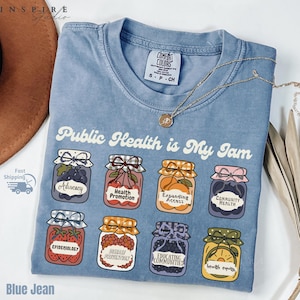 K&ouml;nnte beinhalten: Hellblaues T-Shirt mit dem Aufdruck "Public Health is My Jam" und Illustrationen von Marmeladengl&auml;sern. Jedes Glas zeigt eine andere Frucht und ein Etikett, darunter "Advocacy" und "Epidemiology". Eine goldene Halskette liegt auf dem Shirt.