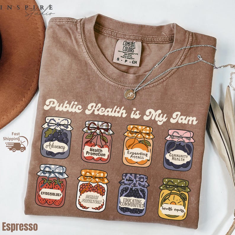 K&ouml;nnte beinhalten: Espressofarbenes T-Shirt mit dem Schriftzug "Public Health is My Jam" und Illustrationen von Einmachgl&auml;sern. Jedes Glas hat ein anderes Etikett, darunter "Advocacy" und "Epidemiology". Eine silberne Halskette mit Anh&auml;nger liegt auf dem Shirt.