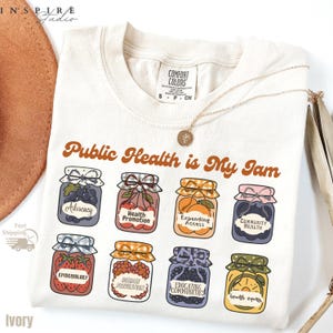 K&ouml;nnte beinhalten: Cremefarbenes T-Shirt mit dem braunen Schriftzug "Public Health is My Jam". Darunter befinden sich Illustrationen von Marmeladengl&auml;sern mit Etiketten wie "Advocacy" und "Epidemiology". Eine goldene Halskette liegt auf dem Shirt.