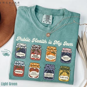 K&ouml;nnte beinhalten: Hellgr&uuml;nes T-Shirt mit dem Schriftzug "Public Health is My Jam" und Illustrationen von Marmeladengl&auml;sern, die jeweils mit Begriffen aus dem &ouml;ffentlichen Gesundheitswesen beschriftet sind. Das T-Shirt hat einen Rundhalsausschnitt und kurze &Auml;rmel.