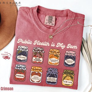 K&ouml;nnte beinhalten: Karmesinrotes T-Shirt mit dem Aufdruck "Public Health is My Jam" und Illustrationen von Marmeladengl&auml;sern, die jeweils mit Konzepten der &ouml;ffentlichen Gesundheit beschriftet sind. Eine silberne Halskette mit Anh&auml;nger liegt auf dem Shirt.