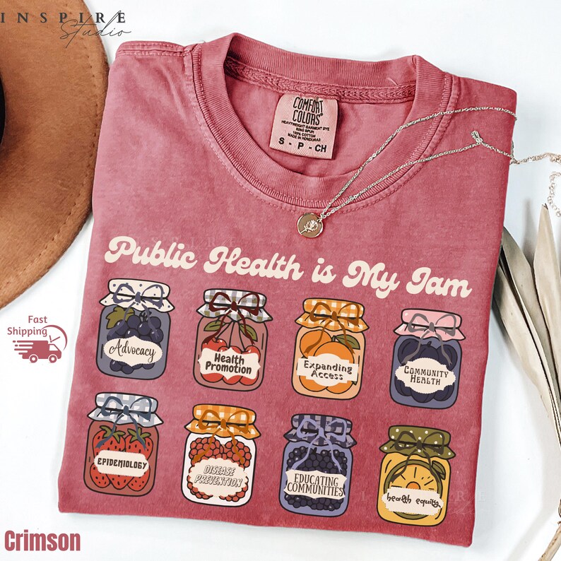 K&ouml;nnte beinhalten: Karmesinrotes T-Shirt mit dem Aufdruck "Public Health is My Jam" und Illustrationen von Marmeladengl&auml;sern, die jeweils mit Konzepten der &ouml;ffentlichen Gesundheit beschriftet sind. Eine silberne Halskette mit Anh&auml;nger liegt auf dem Shirt.