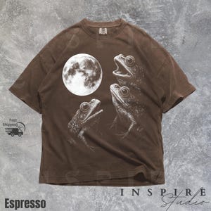 K&ouml;nnte beinhalten: Espressofarbenes T-Shirt mit einer wei&szlig;en Illustration von drei Fr&ouml;schen und einem Vollmond. Die Fr&ouml;sche haben detaillierte Augen und offene M&uuml;nder. Das Shirt hat eine lockere Passform und kurze &Auml;rmel. Der Text "Espresso" befindet sich unten links.