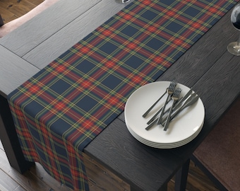 May include: A long rectangular table runner featuring a dark navy blue base with a classic plaid pattern in red, yellow, and green lines. The fabric runner is draped across a dark wooden table surface, accompanied by a stack of white plates and silverware.