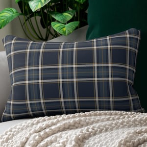 May include: Rectangular pillow with a navy blue plaid pattern. The plaid design features lines in white, light blue, and gold. The pillow is on a white knitted blanket.