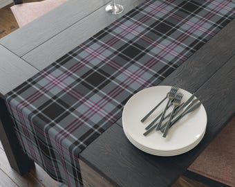 May include: A rectangular table runner featuring a plaid pattern in shades of black, light gray, and thin dark red lines. The runner is laid out on a dark wooden table, with a stack of white ceramic plates and silver forks placed on top.