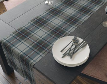 May include: A rectangular table runner featuring a plaid pattern in shades of gray, dark green, and thin red lines. The fabric is draped across a dark wooden table surface, with a stack of white plates and silver forks placed on top of the runner.