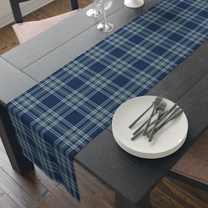 May include: A rectangular table runner featuring a classic plaid pattern in shades of navy blue, light blue, and white. The fabric runner is laid lengthwise across a dark wooden table, providing a decorative textile accent for the dining surface.