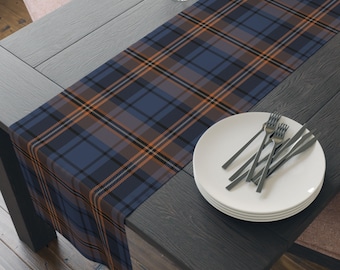 May include: A rectangular table runner featuring a plaid pattern in shades of navy blue, black, and thin orange stripes. The fabric runner is laid flat across a dark wooden table, with a stack of white plates and silver forks placed on one side.