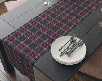 May include: A rectangular table runner featuring a dark gray, white, and deep red plaid pattern. The fabric is laid flat across a dark wooden table surface, with a stack of white plates and silver forks placed on top of the textile runner.