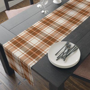 May include: A rectangular table runner featuring a plaid pattern in shades of burnt orange, cream, and light brown. The fabric is draped lengthwise across a dark wooden table, with a stack of white plates and silver forks placed on the surface.