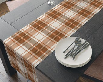 May include: A rectangular table runner featuring a plaid pattern in shades of burnt orange, cream, and light brown. The fabric is draped lengthwise across a dark wooden table, with a stack of white plates and silver forks placed on the surface.