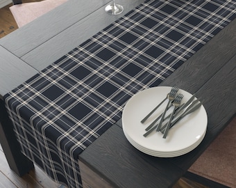 May include: A rectangular table runner featuring a dark navy blue, grey, and white plaid pattern. The runner is laid out lengthwise across a dark wooden table, with a stack of white plates and silver cutlery placed on top of the fabric.