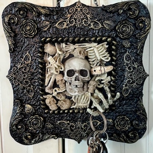 Könnte beinhalten: Ein dekorativer Schlüsselhalter in Schwarz und Gold mit gotischem Thema. Der Rahmen ist mit Rosen und aufwendigen Details verziert. In der Mitte befinden sich ein Schädel und Skelettelemente. Kette und Schlüsselringe sind angebracht.