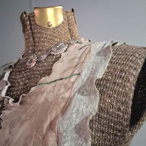 May include: A textured brown mannequin torso wearing a silver-toned concho necklace and layered fabric panels in shades of beige, tan, and muted green. The mannequin features a metallic gold-colored neck plate and a woven, grid-like surface texture.