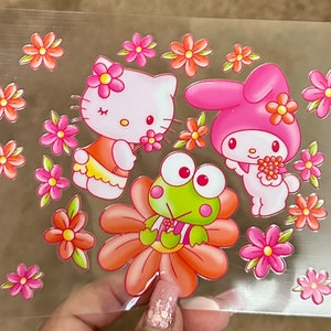 May include: A sheet of clear plastic with colorful stickers featuring Hello Kitty, My Melody, and Keroppi. The stickers are surrounded by pink and orange flowers.