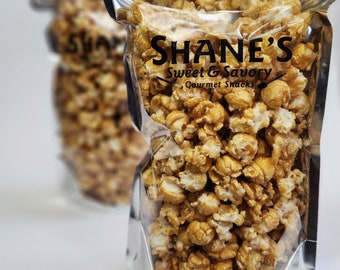 May include: Two clear plastic bags filled with caramel popcorn. The bags have a silver foil top and are sealed. The front bag has the text "Shane's Sweet & Savory Gourmet Snacks" printed in black.