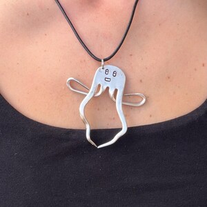 May include: A silver-colored pendant shaped like a ghost, crafted from a repurposed metal fork. The ghost figure features two small eyes and a mouth etched into the top, with curved tines forming arms and a flowing body on a black cord necklace.