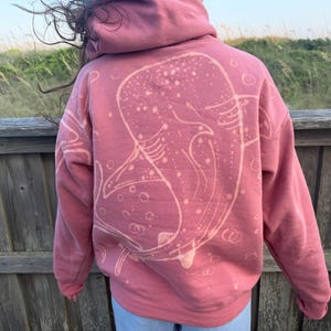 May include: A dusty rose-colored hooded sweatshirt featuring a large, light-colored bleach-style graphic of a whale shark on the back. The design includes circular bubble patterns surrounding the whale shark, with long sleeves and a hood.