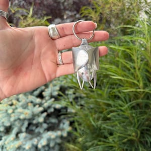 May include: A silver-toned metal keychain crafted from repurposed cutlery, shaped to resemble a bat with folded wings, a rounded head, and two pointed ears at the bottom. The object features a circular split ring at the top for attachment.
