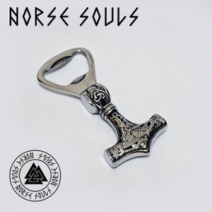 Könnte beinhalten: Ein silberner Flaschenöffner in Form von Thors Hammer, Mjolnir, mit aufwendig gravierten Details. Der Öffner hat einen schwarzen Hintergrund und silberne Akzente. Der Text "NORSE SOULS" befindet sich oben im Bild.